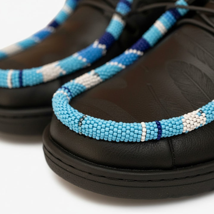 All Nations Designs &amp; More black Hey Dude slip-on shoes featuring blue hand-beaded trim and laser-etched feather patterns.