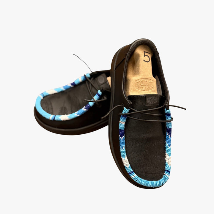 All Nations Designs &amp; More black Hey Dude slip-on shoes with blue hand-beaded trim and laser-etched feather detail.