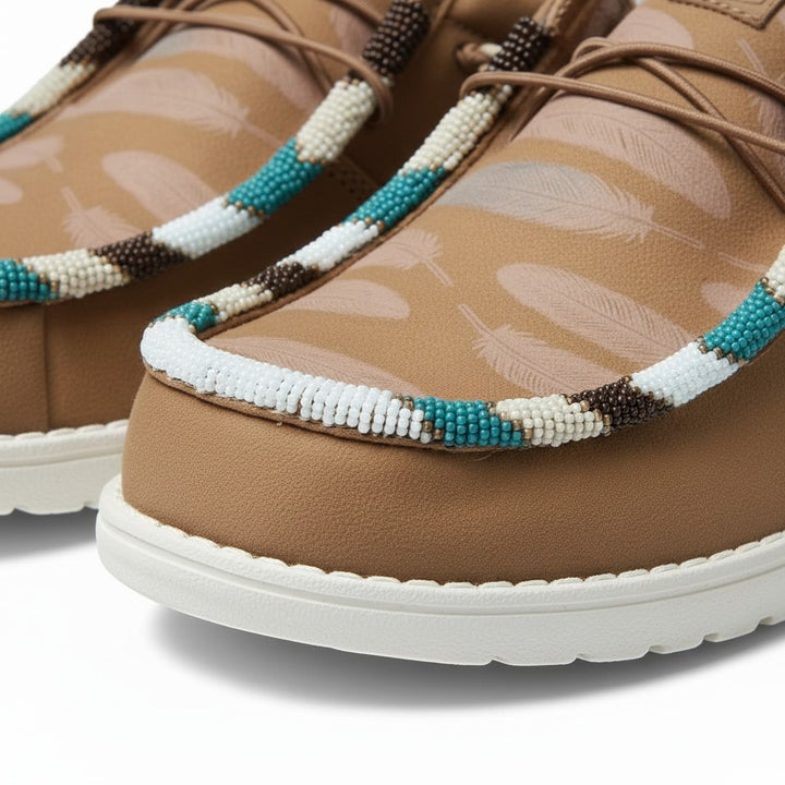 All Nations Designs &amp; More brown Hey Dude slip-on shoes featuring custom hand-beaded feather patterns and white soles.