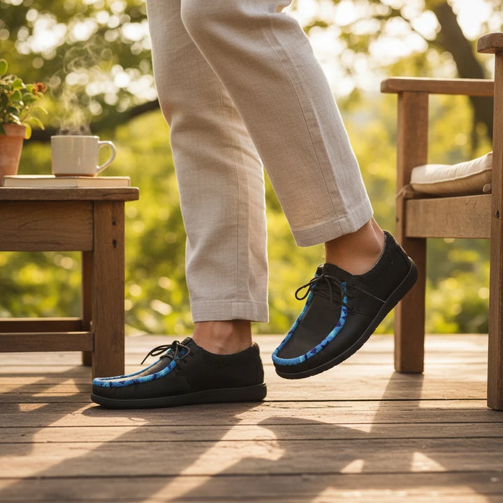 All Nations Designs &amp; More hand-beaded black Hey Dude slip-on shoes with blue feather pattern on a wooden deck