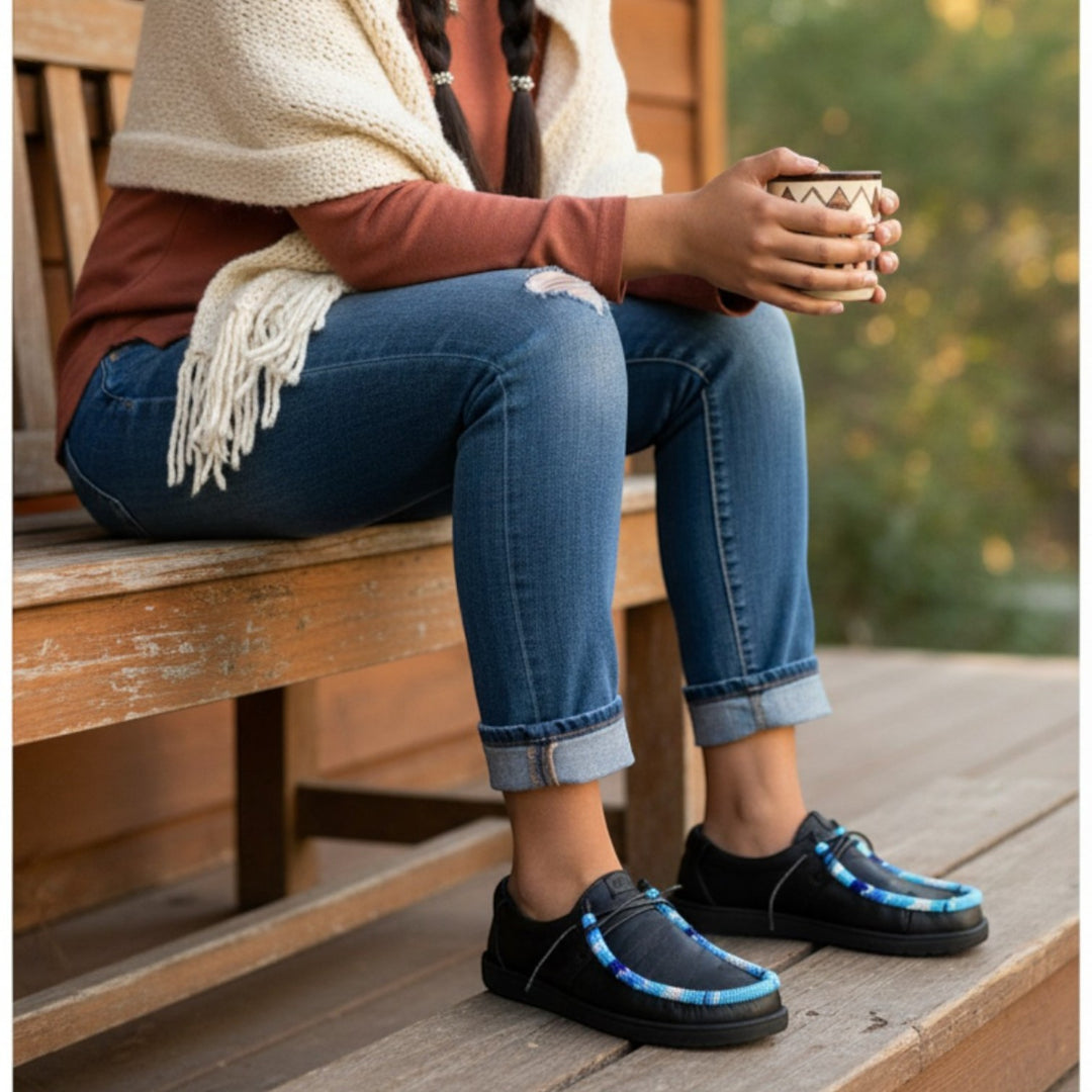 All Nations Designs &amp; More Hey Dude black slip-on shoes with blue hand-beaded detail worn by a woman on a wooden porch.