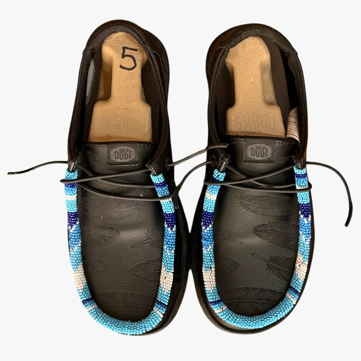 All Nations Designs &amp; More black Hey Dude slip-on shoes featuring laser-etched feathers and blue hand-beaded trim.